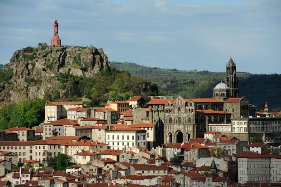 Hilltop town with red tile roofs and a large stone cathedral, with a towering statue on a rocky outcrop above the buildings. Historic destination that can be reached from nearby hiking trails in Europe.