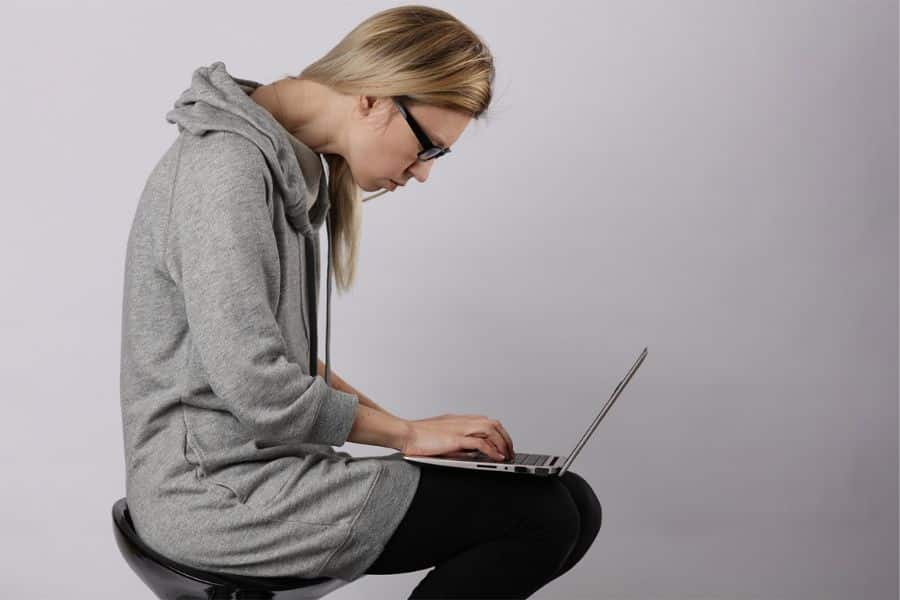 Woman in glasses leans forward with rounded shoulders while typing on a laptop on her lap. Her hunched sitting position shows poor seated posture.