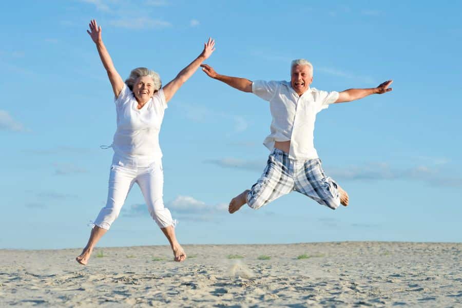 Two older people leaping for joy on the sand under a blue sky