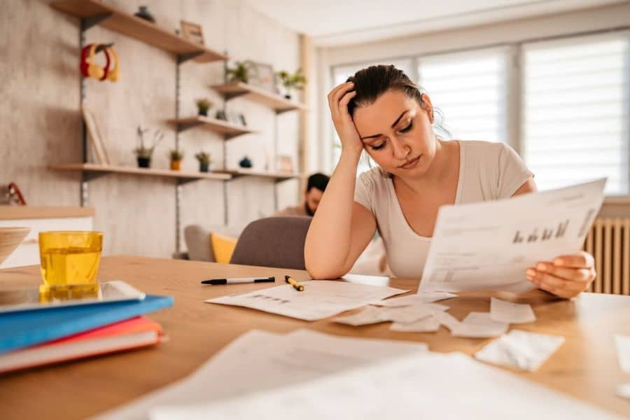 Woman at a table looks stressed while reading a bill with receipts and papers spread out in front of her. The photo shows the worry and pressure of trying to cover monthly expenses.