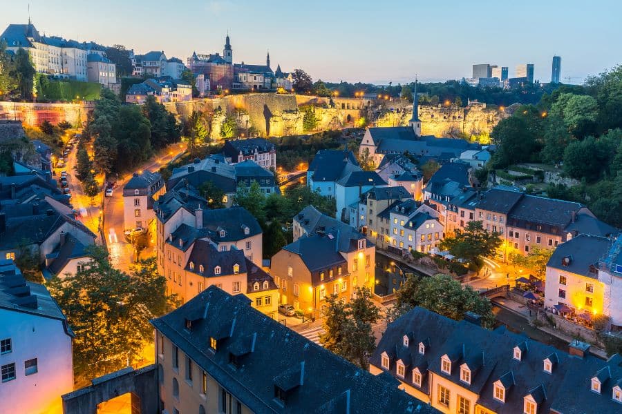 Evening view of a European city with softly glowing streetlights, pastel houses, and a fortress wall lit up on the hill above. The mix of old architecture and twilight light gives the neighborhood a calm welcoming feel.
