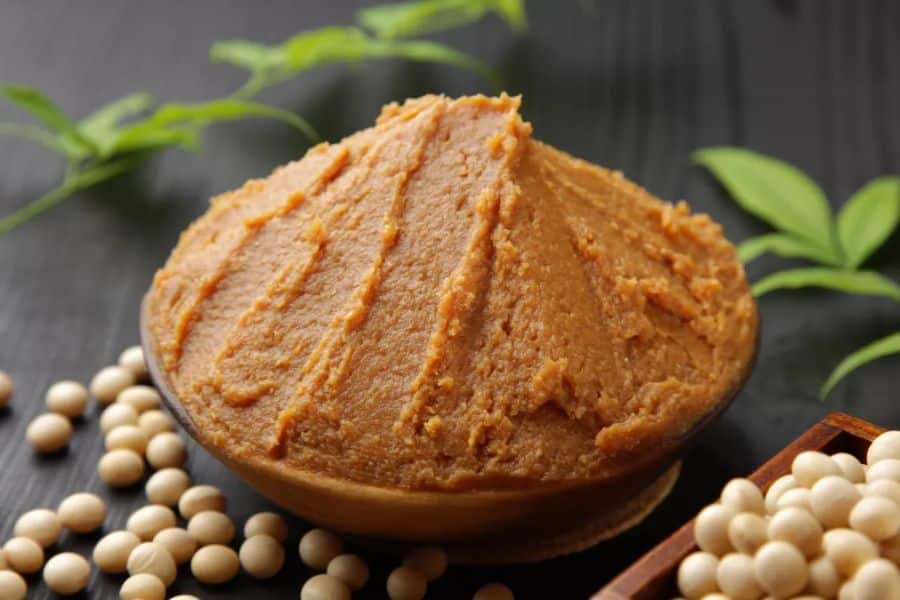 A rounded mound of brown miso paste sits in a wooden bowl with pale soybeans scattered around it. This close up shows a fermented soybean food often associated with gut and brain health.