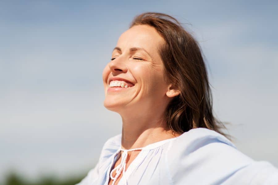 Smiling woman stands outdoors with her eyes closed and face turned toward the sun under a clear blue sky. The scene reflects a calm moment of fresh air and gratitude.