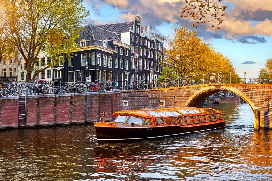 A glass covered tour boat moves through an Amsterdam canal beneath a brick arched bridge lined with parked bicycles and tall narrow houses. The peaceful waterway and walkable setting fit the charm people look for in retiree hotspots in Europe.