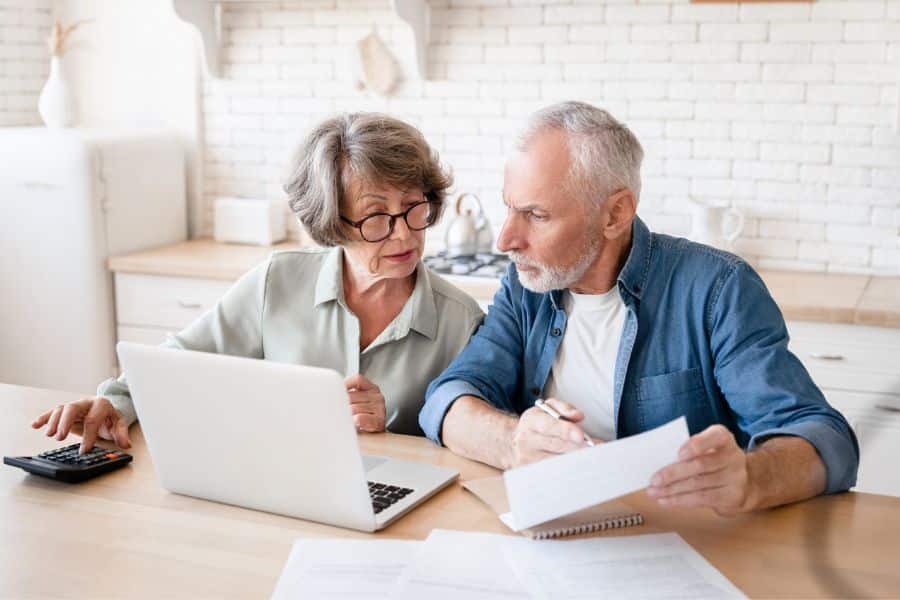 Older couple reviewing paperwork at a kitchen table with a laptop and calculator beside them. The woman points toward the screen while the man studies a document, showing a serious conversation about money and the stock market.