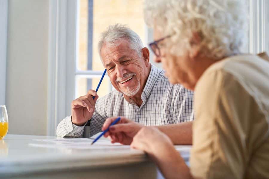 An older couple sit at a table by a bright window, writing on papers and talking together. Planning, reviewing information, and making decisions together reflects brain-boosting habits that keep the mind active.