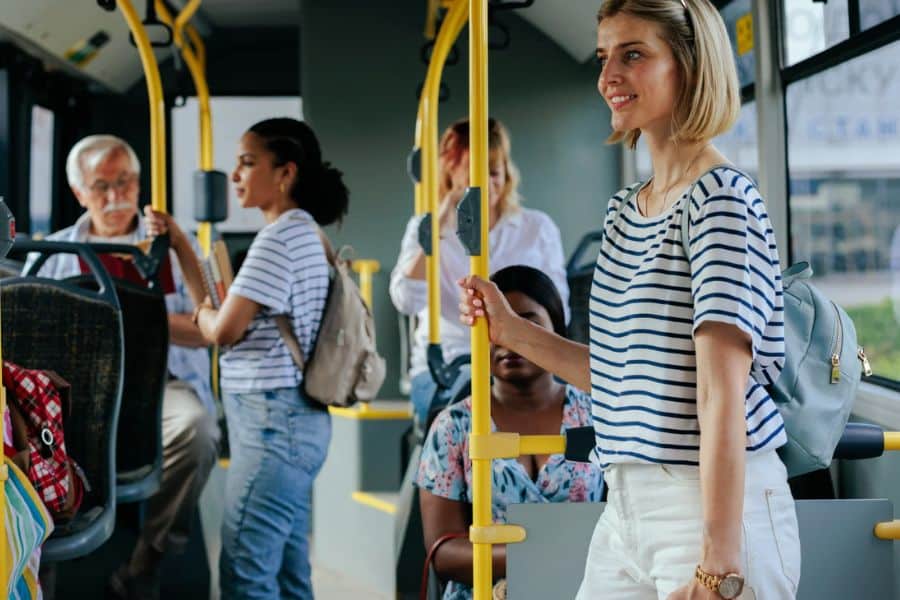 Several passengers stand and sit inside a city bus with yellow handrails and daylight coming through the windows. This everyday public transit scene fits content about inflation and the rising cost of commuting.