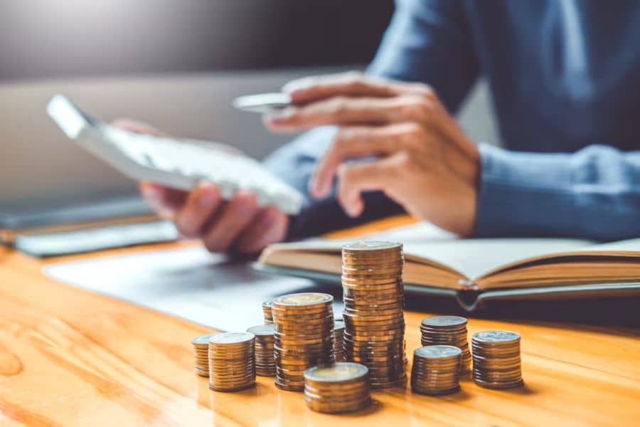 Close up of a person using a calculator with several stacks of coins lined up on a wooden table beside an open notebook. The scene suggests counting savings or tracking stock market related finances.