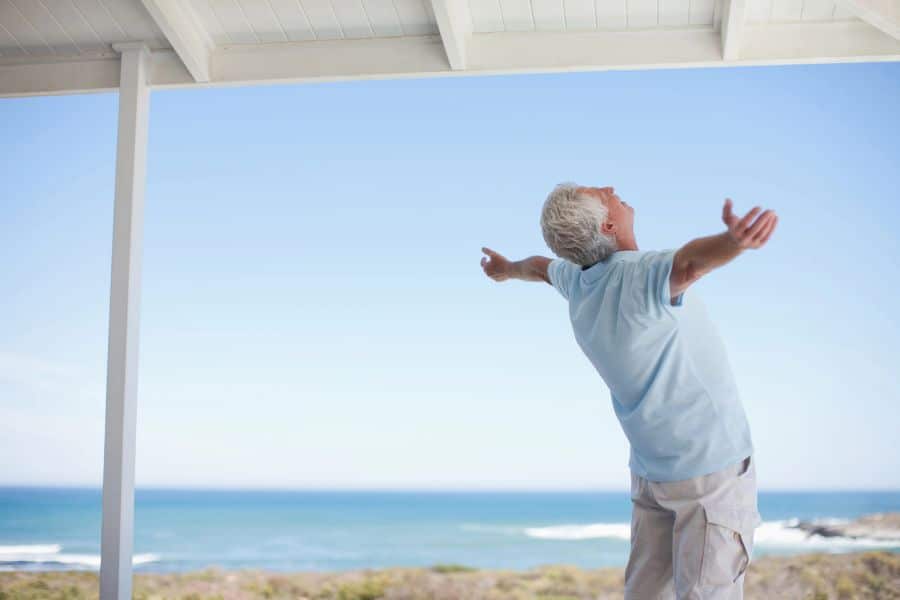 An older man stands on a covered porch facing the ocean with his arms stretched wide toward the sky. The calm coastal setting suggests a mindful moment outdoors that can be part of healthy daily routines.