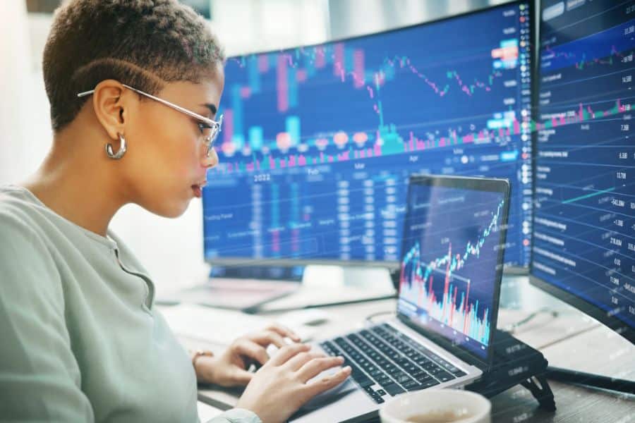 Person wearing glasses works on a laptop in front of large monitors filled with stock charts and market data. The trading setup connects naturally to inflation, investing, and financial decision making.