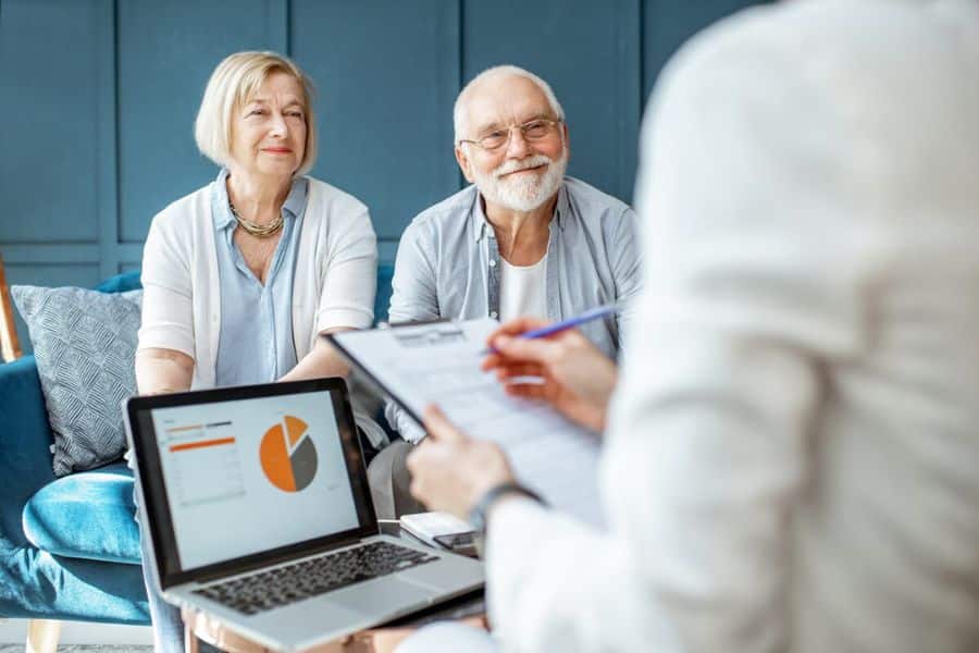 Older couple sitting with a financial advisor while a laptop displays a pie chart and the advisor holds a clipboard in the foreground. Their relaxed expressions suggest a planning meeting about investments or the stock market.