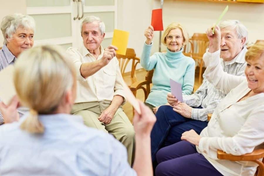 A group of older adults sit in a circle indoors holding up colorful cards during an activity led by a facilitator. Group games and participation like this can be brain-boosting habits that encourage focus, memory, and social interaction.