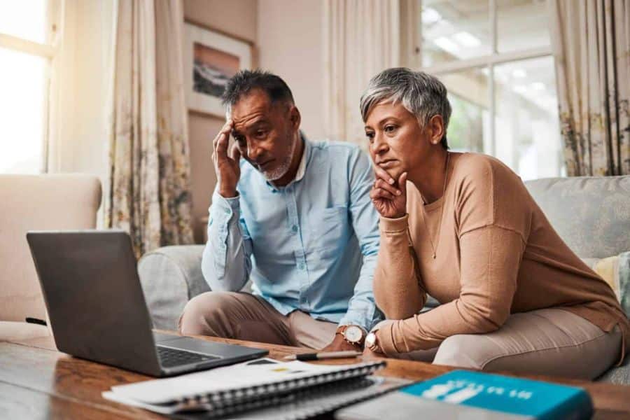 Middle aged couple sitting on a sofa in front of a laptop with concerned expressions as they look at the screen. Papers and a notebook on the table suggest they are reacting to troubling financial or stock market news.