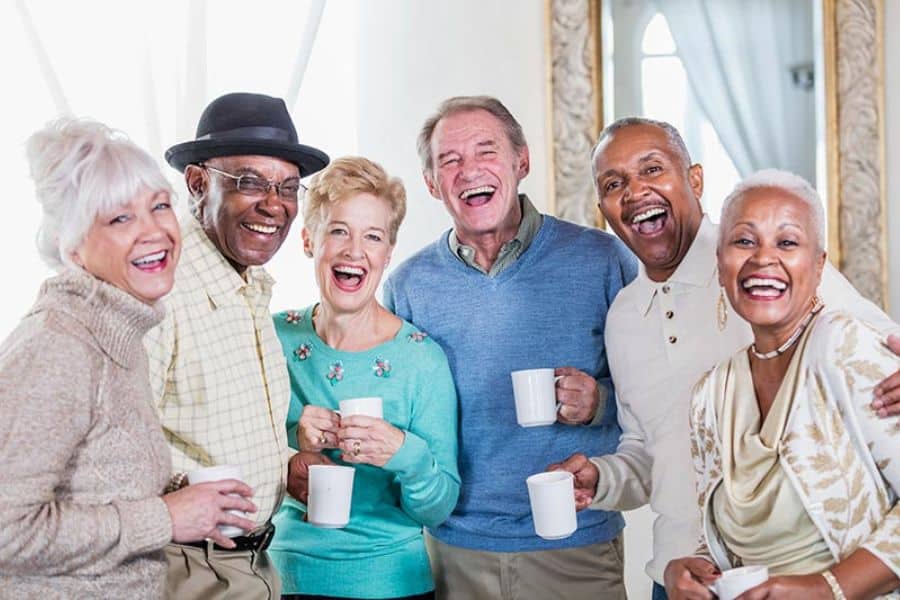 Five older adults stand close together indoors, smiling at the camera and holding white mugs. Their warm, social gathering highlights connection and conversation as part of brain-boosting habits for healthy aging.