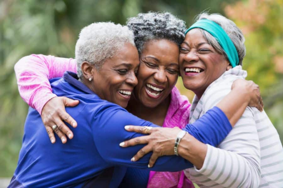 Three women embrace in a group hug outdoors and laugh together. Close friendship and emotional support make this a strong visual for brain-boosting habits built around social connection.