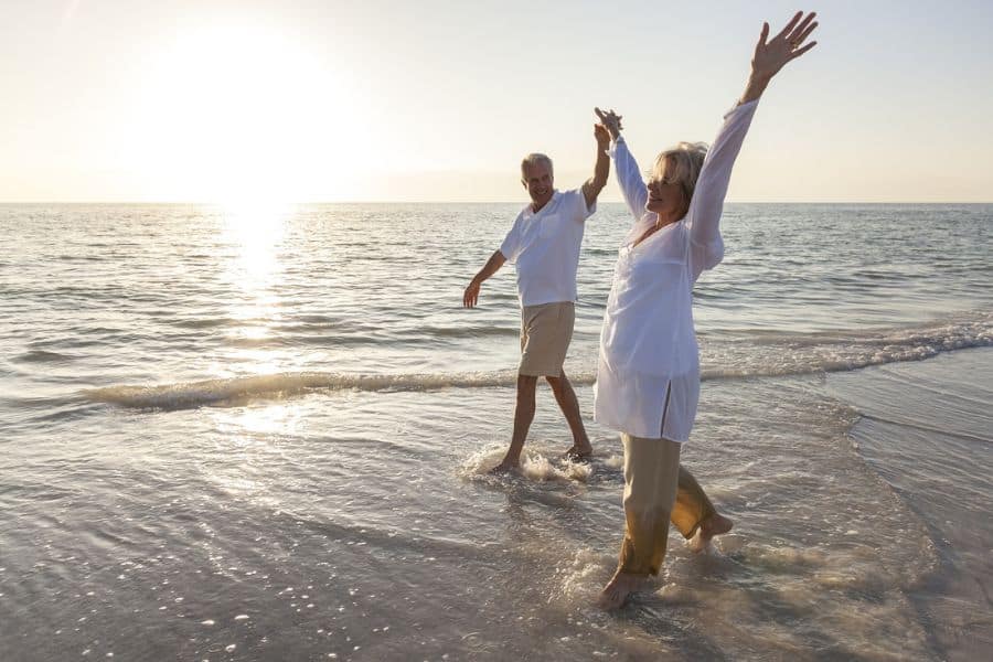 An older couple walk barefoot through shallow ocean water at sunset with their arms raised in celebration. Gentle exercise in nature captures brain-boosting habits that support both mood and mental wellness.