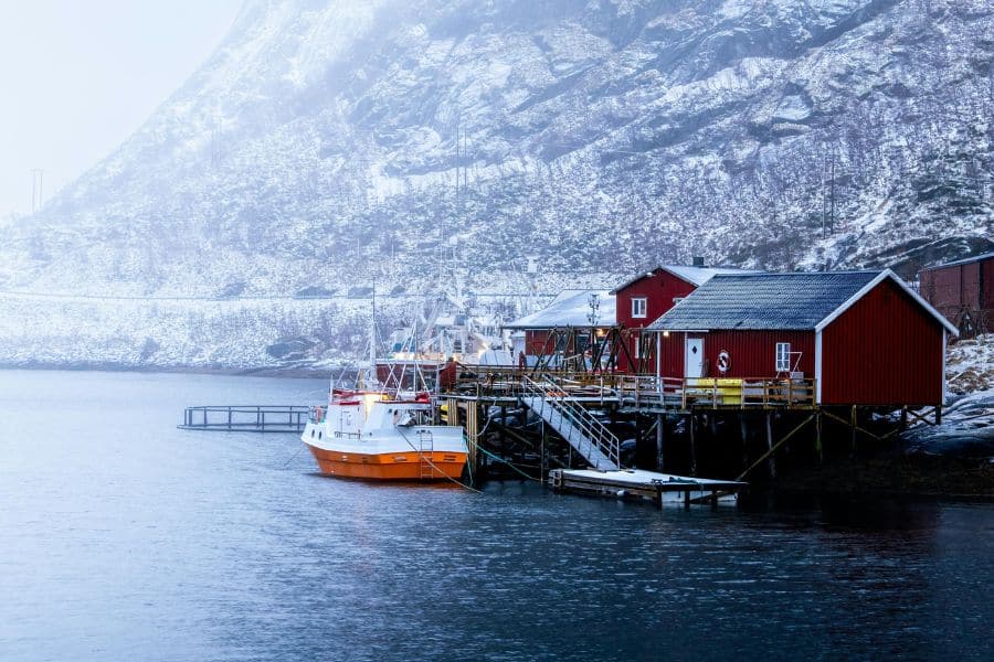 Red wooden cabins and a small fishing boat sit beside still dark water with snowy mountains rising behind them in misty light. The quiet harbor scene feels cold, remote, and serene.