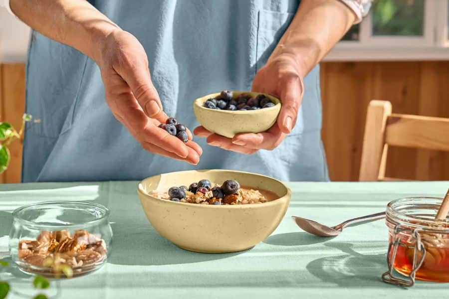 Hands sprinkle fresh blueberries over a bowl of oatmeal topped with nuts on a sunlit table. This balanced breakfast contrasts with the kind of morning rituals destroying your metabolism discussed in the article.