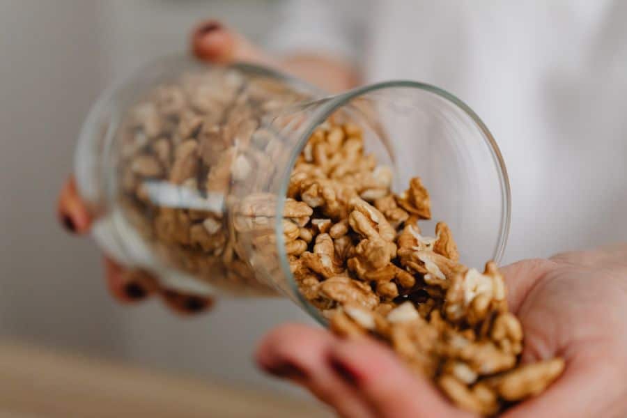 Walnut halves pour from a clear glass jar into an open hand, with the background softly blurred. The close up view emphasizes nuts as everyday foods that are easy to store and grab for cooking or snacks.