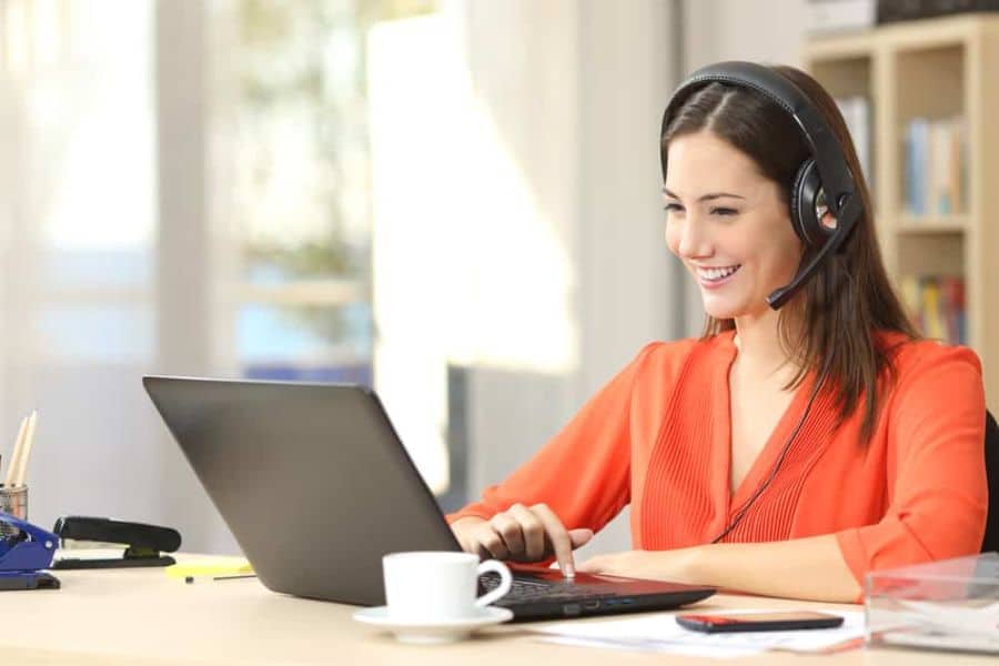 Woman wearing a headset smiles at her laptop while working at a desk with a coffee cup nearby. This is a strong visual for work from home jobs involving customer service virtual support or online communication.