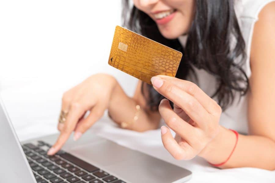 Woman uses a laptop while holding a gold credit card in front of a plain light background. This supports content about online spending habits and ways to save money when shopping with a credit card.
