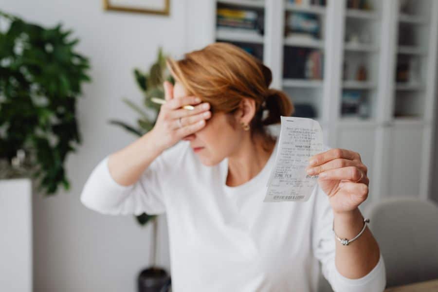 Woman covers part of her face with one hand while holding up a receipt in the other in a bright home office. The moment reflects stress after seeing a purchase total or household cost.