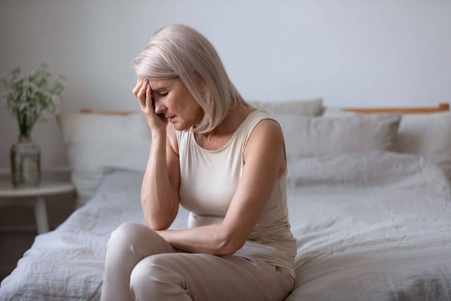 Older woman sits on the edge of a bed with her head in her hand, looking fatigued and distressed in a quiet bedroom. The photo conveys sudden weakness or discomfort that can overlap with stroke symptoms.
