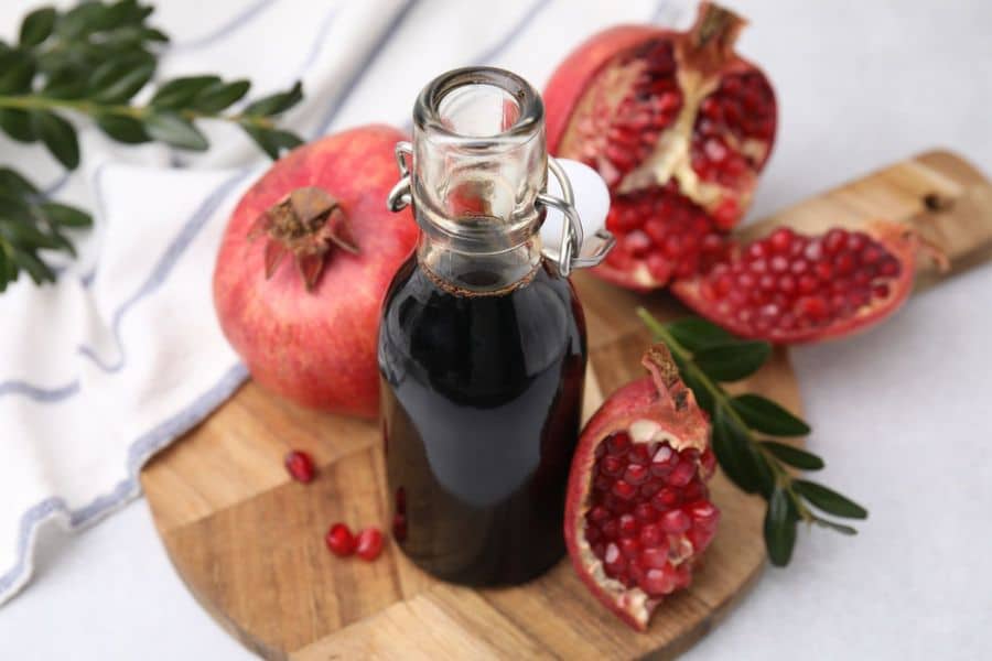 Glass bottle of dark pomegranate molasses stands on a wooden board surrounded by whole and sliced pomegranates. The glossy red seeds and deep syrup emphasize the fresh fruit and concentrated sauce.