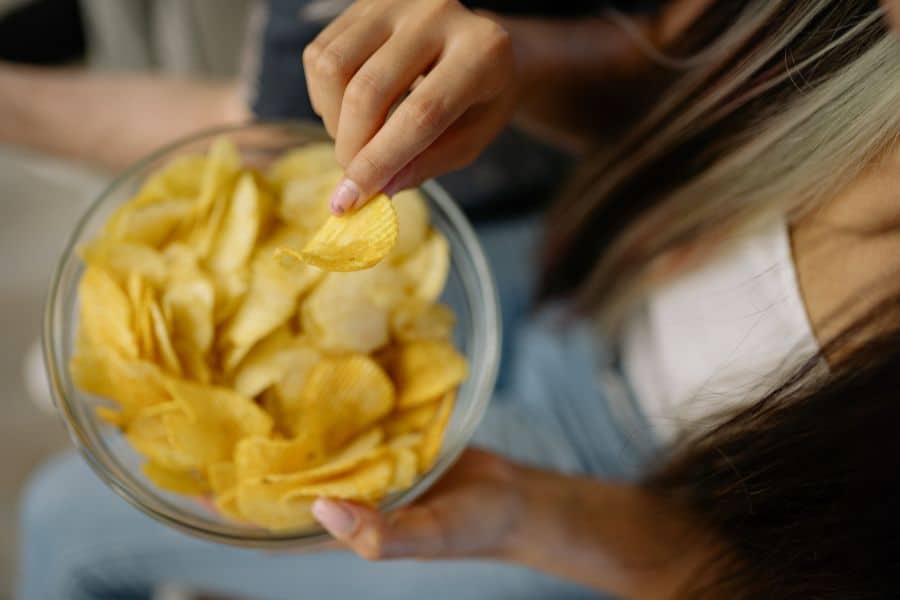 Person holding a glass bowl of ridged potato chips while reaching in to grab one chip. This close view of snacking can support content about mindless eating and hunger hormones.