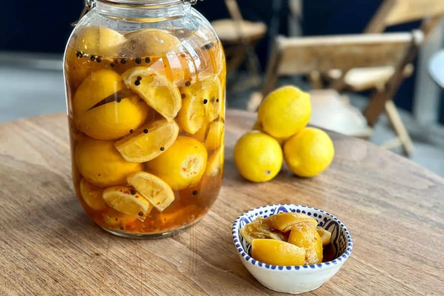 Large glass jar filled with preserved lemons in brine sits on a wooden table with a small bowl of lemon pieces in front. Whole lemons in the background help show the ingredient before and after preserving.