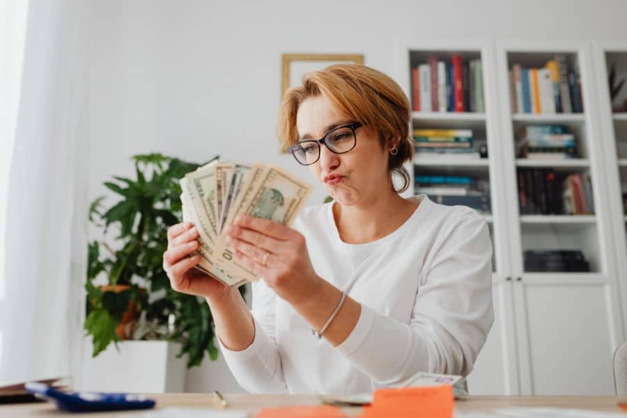 Woman in glasses sits at a desk at home counting a fan of cash bills with bookshelves and a plant behind her. Her expression suggests she is checking savings or comparing expenses.