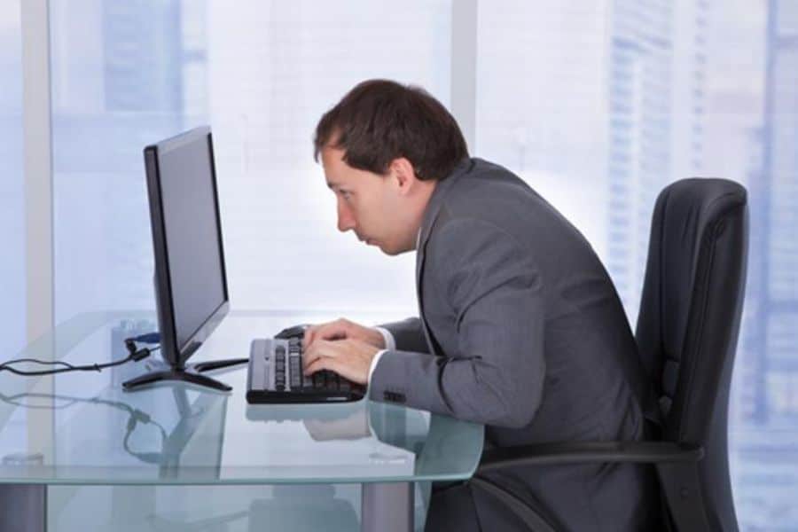 Man in a suit leans very close to a desktop monitor while typing at a glass desk. His cramped sitting position shows poor office posture.