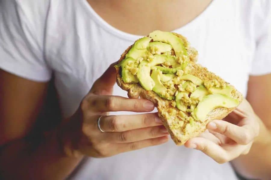 Person in a white shirt holds a slice of toast topped with avocado slices and mashed avocado. This shows a simple avocado meal that fits naturally into content about ways to lower your cholesterol.