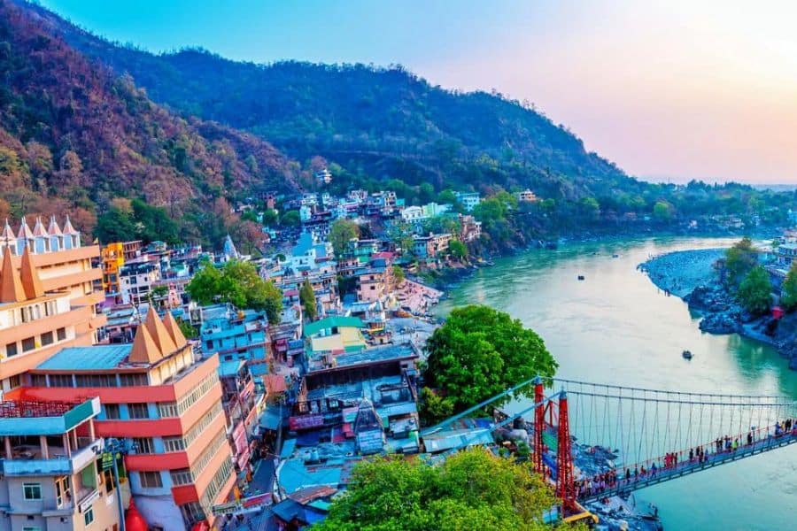 A river runs through a busy town surrounded by forested hills, with a suspension bridge crossing the water and colorful buildings packed along the bank. This suits vacation destinations because it combines mountain scenery, local architecture, and a lively riverside setting.