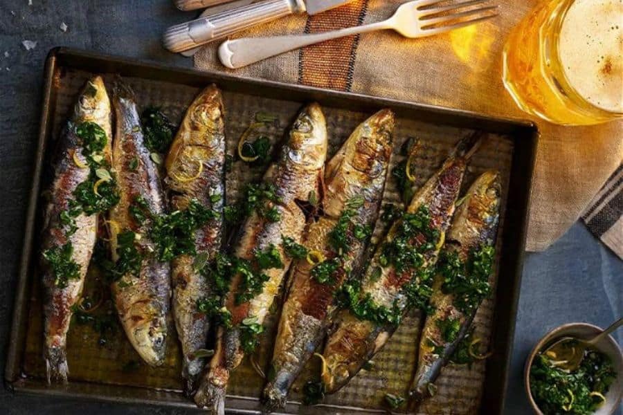 Roasted whole fish are lined up in a dark baking tray and finished with chopped green herbs and lemon slices. A glass of beer and serving utensils sit nearby, giving the scene a rustic dinner table feel.