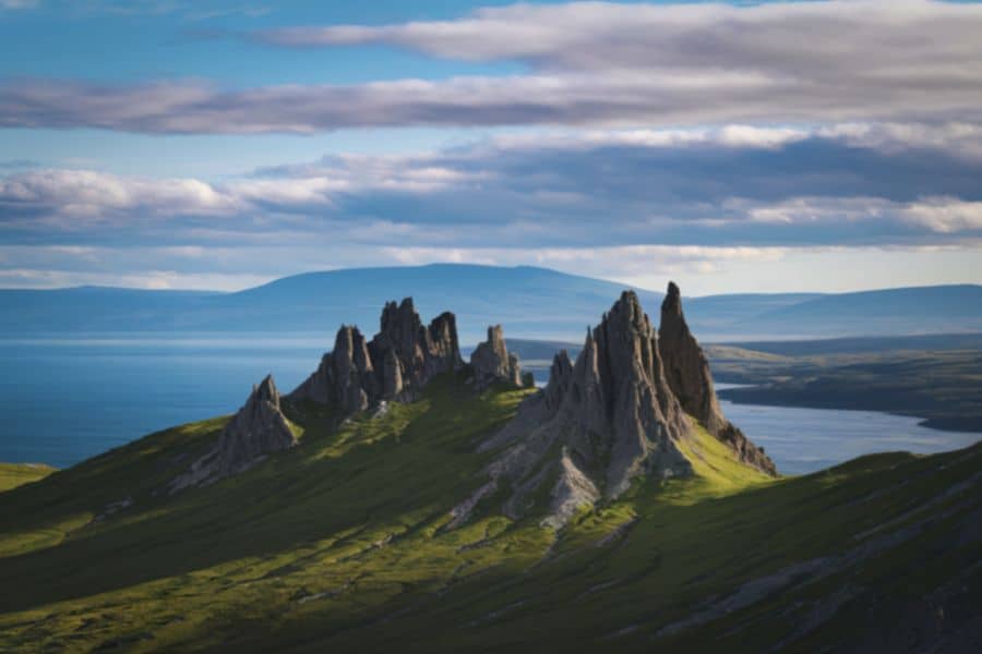 Sharp rocky spires rise from rolling green hills beside a wide stretch of water under a sky layered with soft gray clouds. Light and shadow across the landscape emphasize the rugged natural beauty of this northern coastal view.