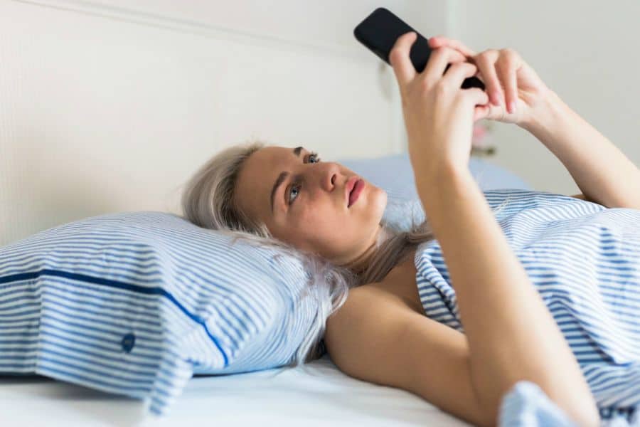 A woman lies in bed under a striped blanket while looking at her phone. The image suggests scrolling on a screen first thing in the morning.