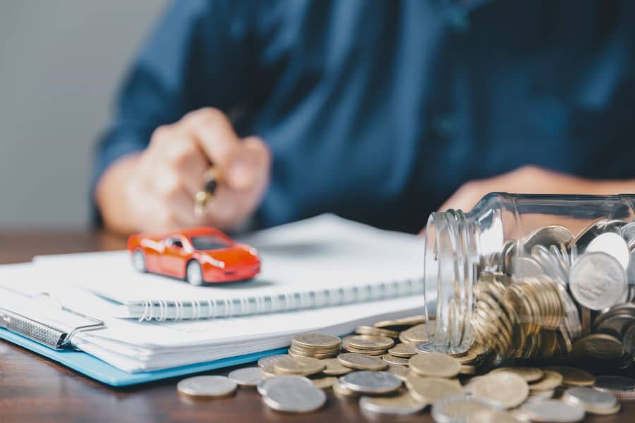 Glass jar tipped over with coins spilling across a desk beside a small red toy car and paperwork in the background. The scene suggests setting aside money for vehicle costs or finding ways to save money on insurance.