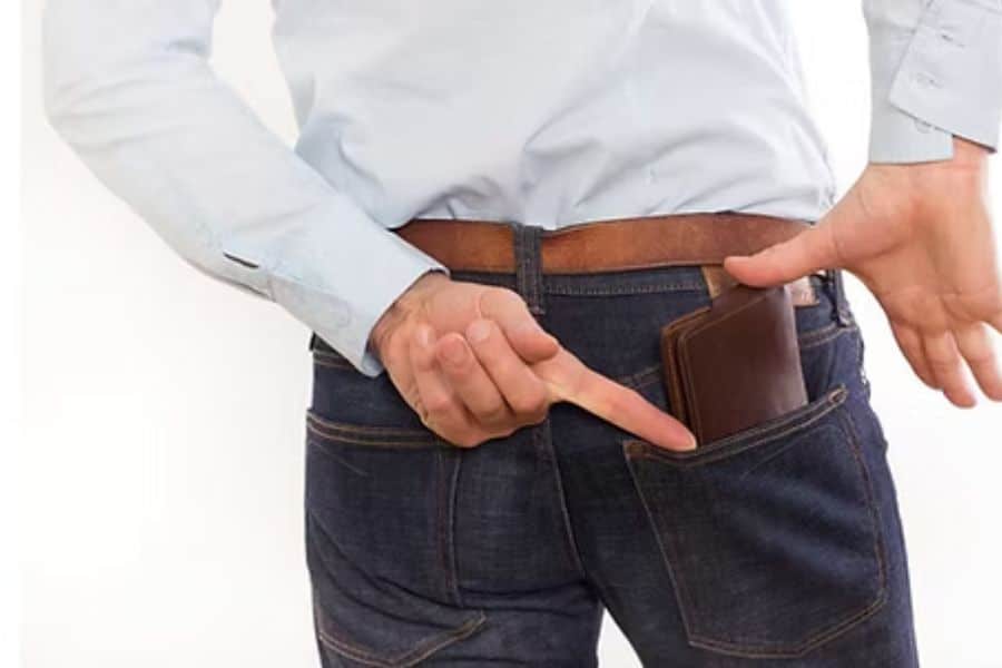 Close view of a person standing with a wallet tucked into a back jeans pocket while pointing at it. This visual relates to sitting position because bulky back pockets can affect seated comfort and posture.