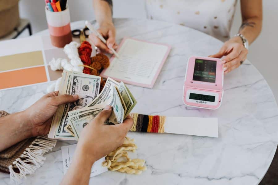 Hands count U.S. cash across a marble table during a kakeibo budgeting session while another person writes in a notebook beside a pink calculator. Yarn samples and color swatches on the table suggest planning purchases carefully and tracking spending by category.