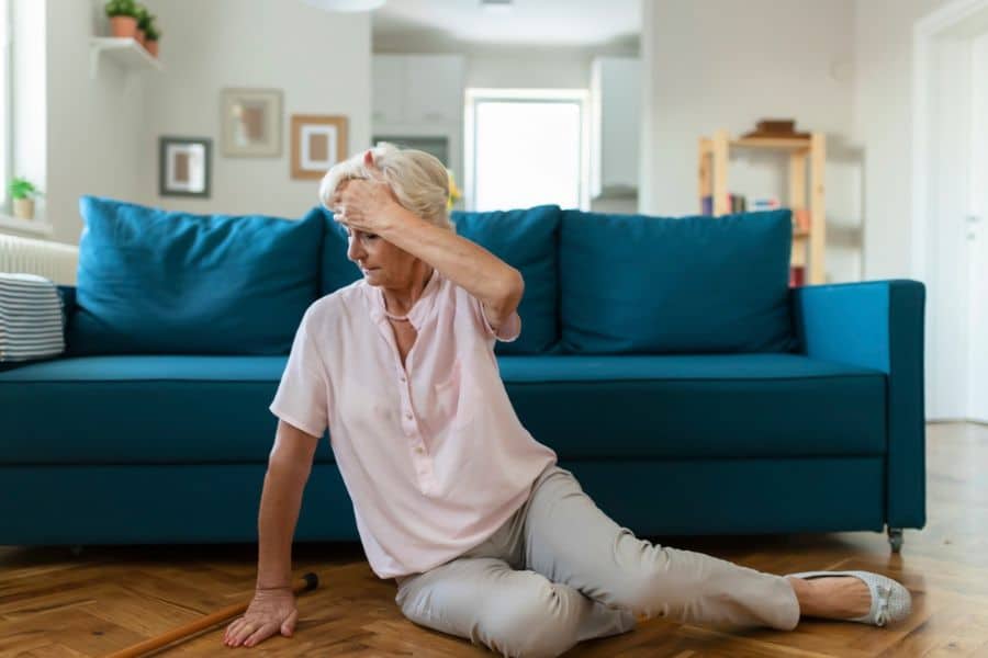 Senior woman sits on a wood floor in front of a blue couch with one hand bracing herself and the other touching her forehead. Her unstable seated position suggests she may have become weak or dizzy from possible stroke symptoms.