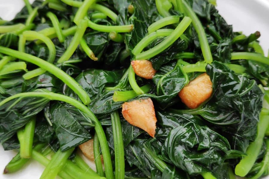 Cooked leafy greens with thick stems are served with pieces of browned garlic on a white plate. This is a strong visual for edible leaves because it shows greens prepared as a simple vegetable side dish.