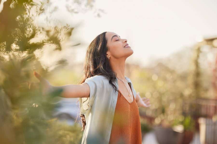 Woman stands outside with her arms open and eyes closed in warm golden light surrounded by soft greenery. This could illustrate a micro habit like stepping outside for a few deep breaths.