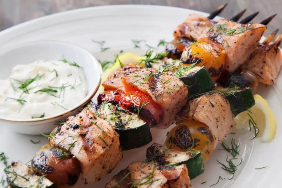 Grilled salmon and vegetable skewers are plated with a bowl of white yogurt sauce and lemon wedges. Char marks on the salmon and the fresh dill on top make the dish look bright and savory.