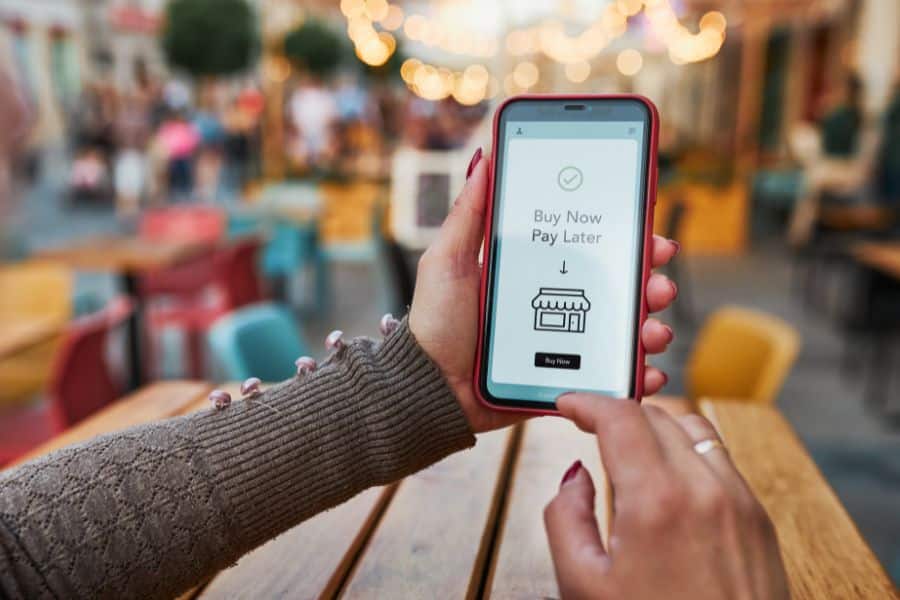 Hand holds a smartphone displaying the text "Buy Now Pay Later" above a store icon and a "Buy Now" button in a blurred café setting. This is useful for content about save money choices because it highlights a payment option that can affect budgeting and debt.