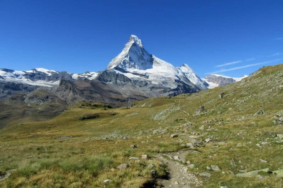 Narrow dirt path winds through alpine grassland toward a jagged snow covered peak under a clear blue sky. High mountain scenery typical of hiking trails in Europe.