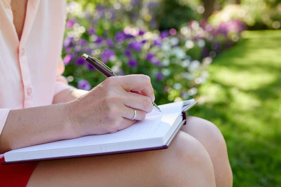 A person writes in a notebook while sitting outdoors in a garden with purple and white flowers blurred in the background. The calm setting fits a reflective kakeibo habit of journaling expenses and money goals by hand.