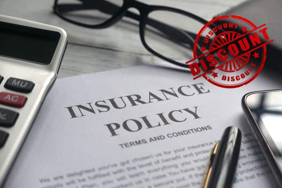 Close view of a document labeled "INSURANCE POLICY" and "TERMS AND CONDITIONS" with a red stamp that reads "DISCOUNT" repeated around the seal and across the center. A calculator glasses phone and pen surround the paper to reinforce the idea of comparing policies and discounts.