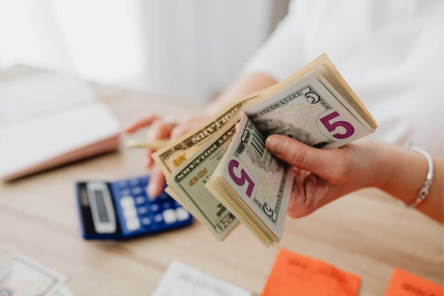 A hand holds several U.S. bills above a desk with a calculator papers and sticky notes during a kakeibo budgeting check in. The close up focuses on cash handling and simple tools used to review spending and savings.