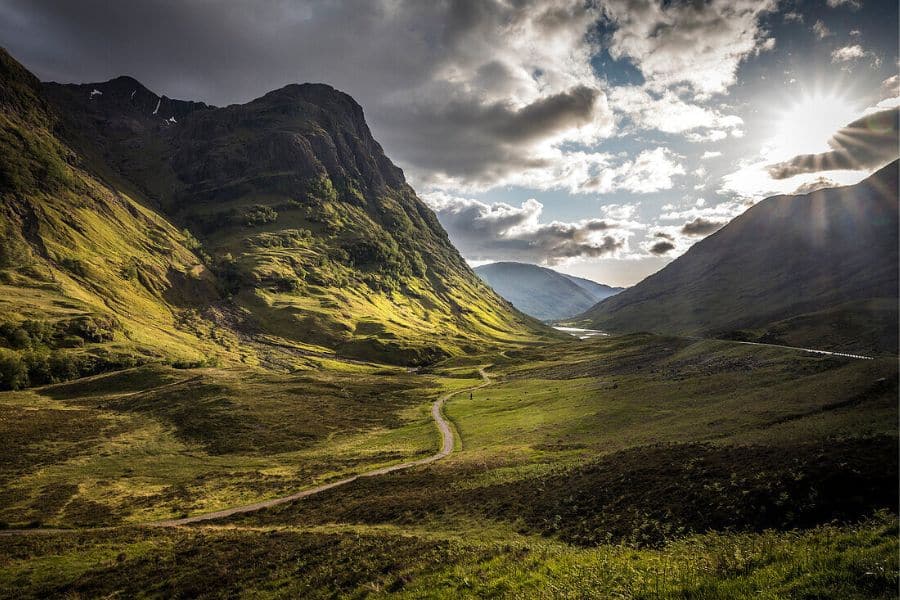 Sunlight breaks through dramatic clouds over a wide green valley between steep dark mountains, with a thin road or trail cutting through the landscape. Moody mountain view that matches hiking trails in Europe.